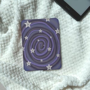 May include: A rectangular e-reader cover featuring a dark purple and light purple spiral pattern across the surface. The design is decorated with multiple white five-pointed stars scattered throughout the swirling vortex graphic on the front.