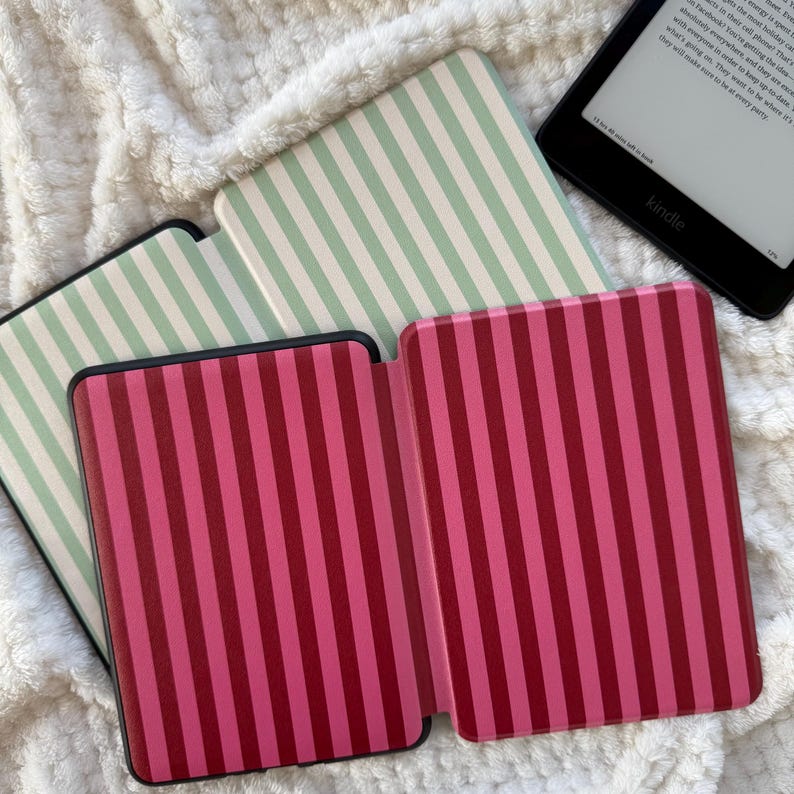 May include: A collection of Kindle covers with vertical stripes in shades of pink and mint green. One cover is open, revealing the interior. A Kindle e-reader is visible in the background, displaying text on its screen.