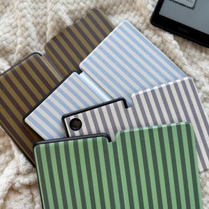 Könnte beinhalten: Eine Auswahl an Kindle-Hüllen in verschiedenen Farben, darunter Grün, Blau und Braun, jeweils mit einem Streifenmuster. Die Hüllen sind auf einer weichen, strukturierten Oberfläche angeordnet, mit einem Kindle-Gerät im Hintergrund.