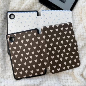 May include: Two e-reader cases are displayed. One case features a dark brown exterior with a repeating pattern of small white hearts. The other case is open, revealing a light cream interior with a delicate blue floral print and a circular cutout.