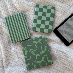 Peut inclure: Trois étuis pour Kindle avec différents motifs : rayures verticales, damier et nœuds. Les étuis sont dans des tons de vert et sont présentés sur une surface blanche texturée. Un lecteur électronique est également visible.