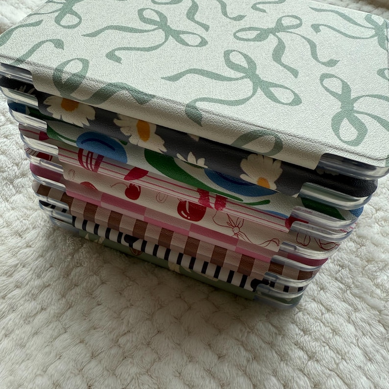 Puede incluir: Una pila de cuadernos decorativos con varias cubiertas estampadas. El cuaderno superior presenta un fondo verde claro con un dise&ntilde;o de lazo repetido. Otras cubiertas muestran patrones florales, de cerezas y de rayas. Los cuadernos se mantienen unidos con pesta&ntilde;as de pl&aacute;stico transparente.
