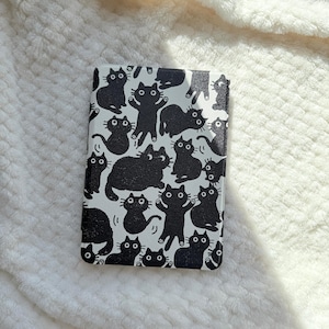 May include: A rectangular e-reader case featuring a repeating pattern of black cats on a white background. The cats are depicted in various poses, including sitting, standing, and reaching, all with wide eyes and simple, rounded features.