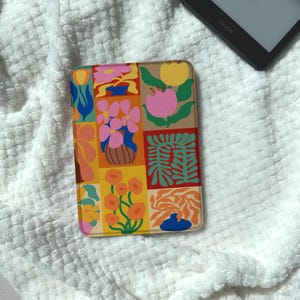 May include: A colorful Kindle case featuring a patchwork design of flowers and abstract shapes in pink, orange, blue, and green. The case is rectangular and has rounded corners. A Kindle e-reader is partially visible in the background.