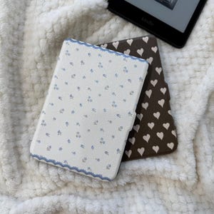 May include: Two e-reader cases are shown. The top case is cream-coloured with a small blue floral pattern and a scalloped blue border. The bottom case is dark brown with a repeating pattern of small, light-coloured hearts. Both are rectangular.