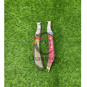 May include: Two hoof knives with stainless steel blades and colorful wooden handles. One handle has a red and black design, the other has a green, pink, and black design. The knives are on a green surface.