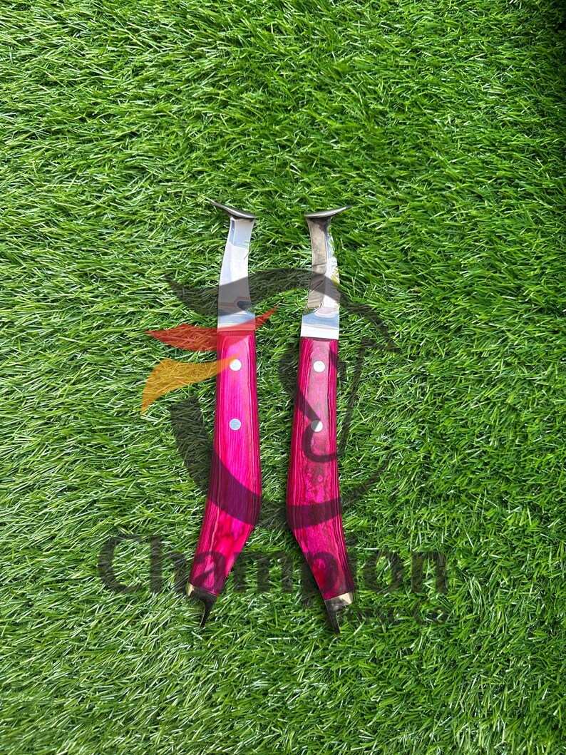 May include: Two knives with bright pink wooden handles and silver blades are displayed against a green background. The handles have a dark wood grain pattern and are accented with brass details. The blades are curved.