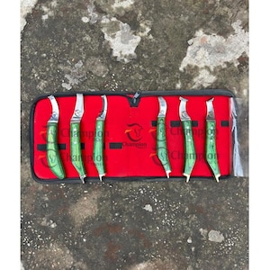 May include: A set of seven hoof knives with green wooden handles and silver blades, displayed in a red case. The case has the word "Champion" printed on it.