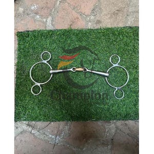 May include: A stainless steel horse bit with a central joint and rounded rings. The bit features a wooden centre piece and is designed for equestrian use. The bit is set against a green background.