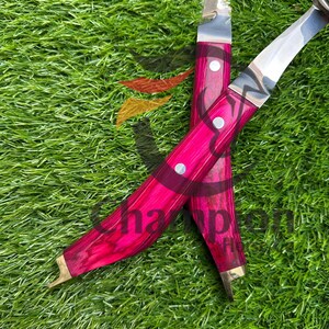 May include: Two curved knives with bright pink wooden handles and silver blades are displayed on green grass. The handles are secured with silver rivets. The blades have a curved shape.
