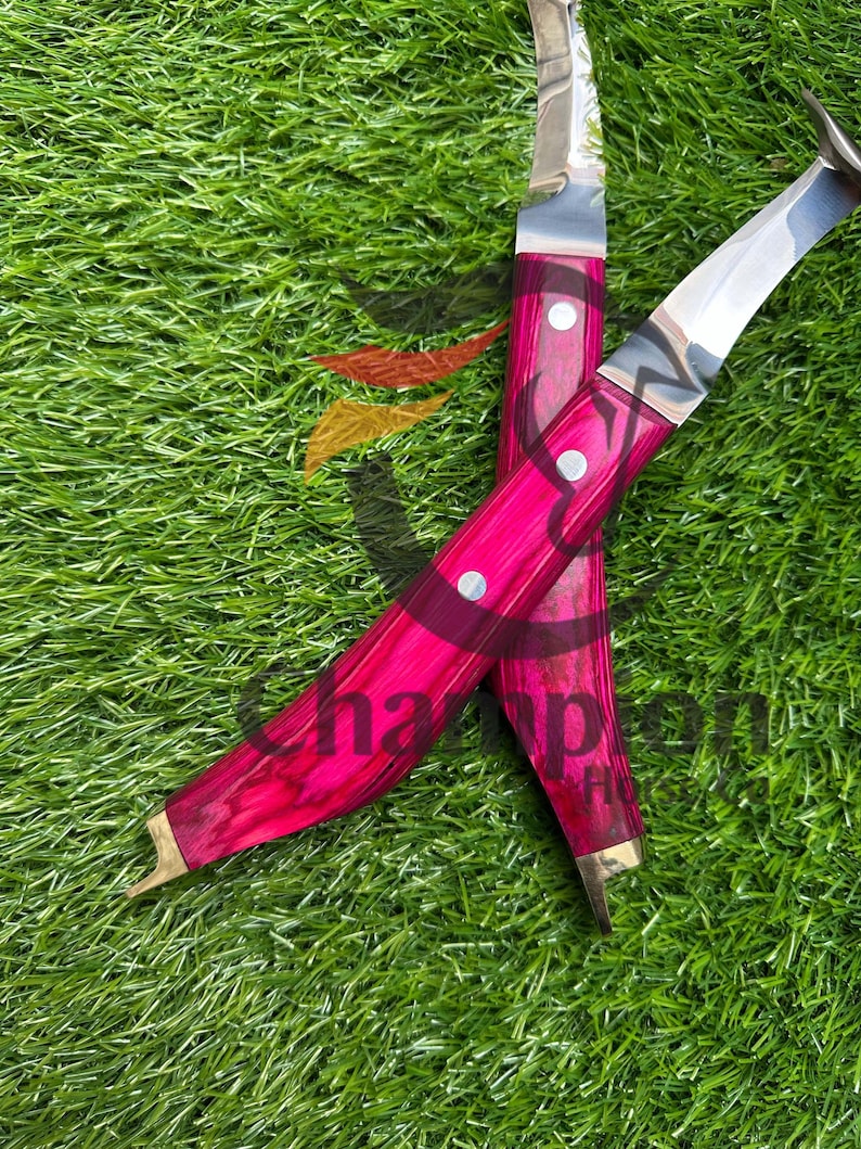 May include: Two curved knives with bright pink wooden handles and silver blades are displayed on green grass. The handles are secured with silver rivets. The blades have a curved shape.