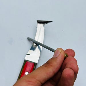 Pu&ograve; includere: Un affilacoltelli in uso su un coltello. Il coltello ha un manico rosso, verde e marrone. L'affilatore &egrave; un'asta grigia con un'estremit&agrave; dorata. La lama del coltello &egrave; argentata.