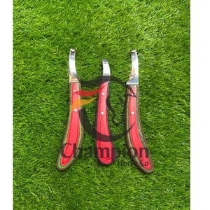 May include: Three hoof knives with red and wood grain handles and curved stainless steel blades. The handles have white dot accents. The knives are arranged on a bed of green grass.