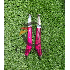 May include: Two knives with bright pink handles and silver blades are displayed on green grass. The handles have a wooden grain appearance and are accented with silver rivets. The blades are curved and have a sharp edge.