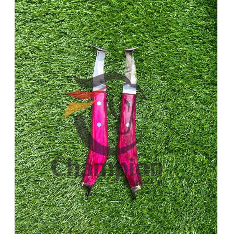 May include: Two knives with bright pink handles and silver blades are displayed on green grass. The handles have a wooden grain appearance and are accented with silver rivets. The blades are curved and have a sharp edge.