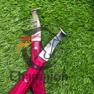 May include: Two hoof knives with bright pink wooden handles and silver blades are crossed on green grass. The handles have a wood grain pattern and are secured with silver rivets.