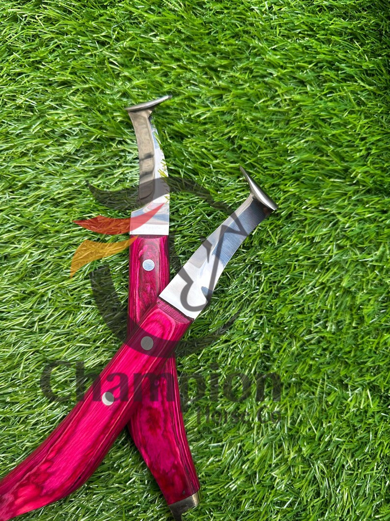 May include: Two hoof knives with bright pink wooden handles and silver blades are crossed on green grass. The handles have a wood grain pattern and are secured with silver rivets.