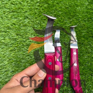 May include: Three hoof knives with polished silver blades and vibrant pink wooden handles. The handles are secured with silver rivets. The knives are displayed against a backdrop of green grass.