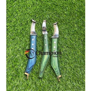 Puede incluir: Tres cuchillos para cascos con mangos de madera en tonos azules y verdes, con hojas plateadas y detalles en latón. Los cuchillos están colocados sobre un lecho de hierba verde. El texto "Champion Horse Co." es visible.
