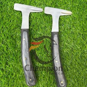 Peut inclure: Deux marteaux en métal avec une tête en forme de T et un manche en bois gris foncé. Les manches ont des rivets en laiton. Les marteaux sont sur une surface verte.