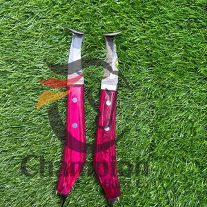 May include: Two knives with bright pink handles and silver blades are displayed on green grass. The handles have a textured appearance and are secured with silver rivets. The blades have a curved shape.