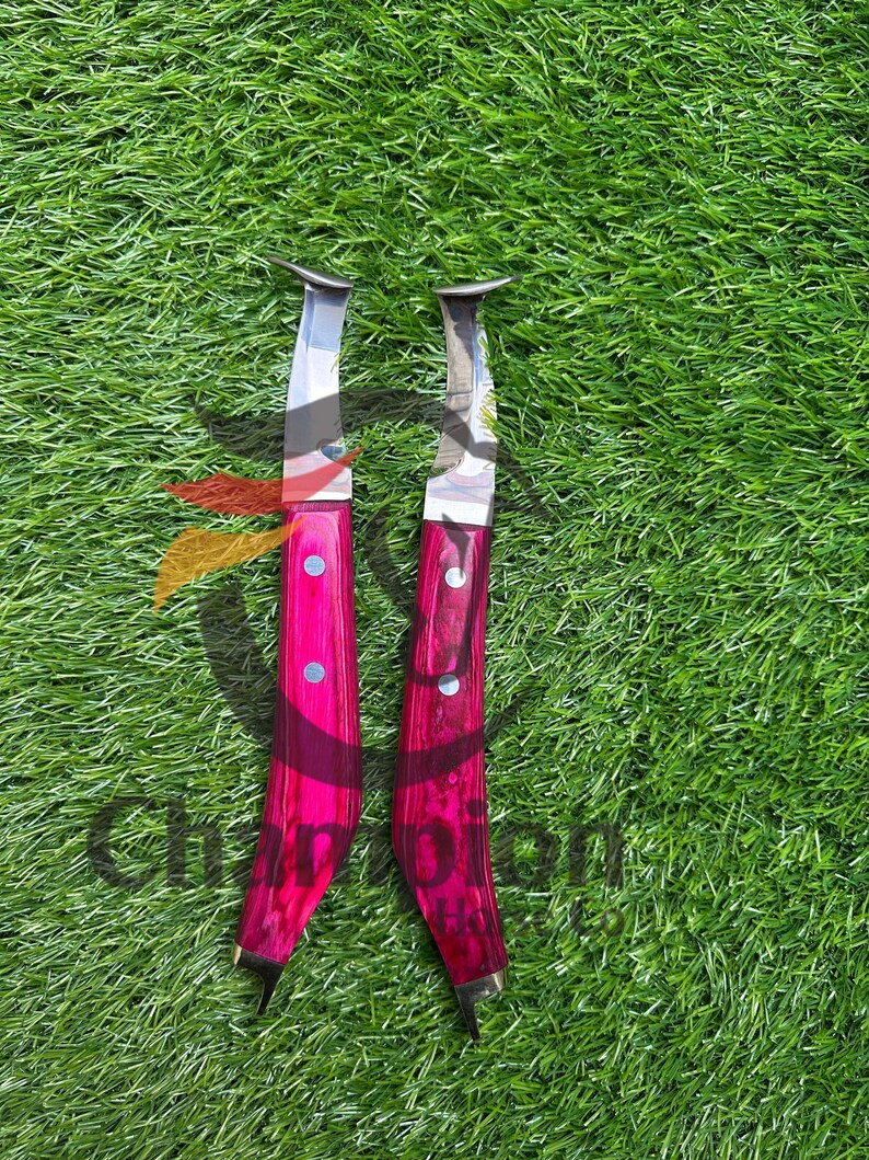 May include: Two knives with bright pink handles and silver blades are displayed on green grass. The handles have a textured appearance and are secured with silver rivets. The blades have a curved shape.