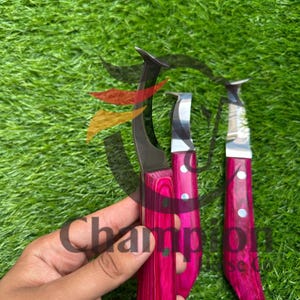 May include: Three hoof knives with curved blades and vibrant pink wooden handles. The blades are silver, and the handles feature a wood grain pattern. The word "Champion" is visible on the image.