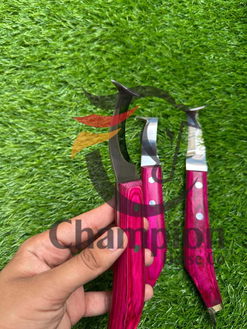 May include: Three hoof knives with curved blades and vibrant pink wooden handles. The blades are silver, and the handles feature a wood grain pattern. The word "Champion" is visible on the image.