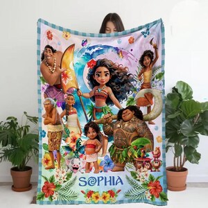 May include: A plush blanket featuring characters from the animated film Moana. The design includes Moana, Maui, and other characters, with the name "SOPHIA" in a stylized font. The blanket has a blue and white checkered border and floral accents.