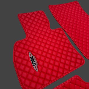 May include: Set of bright red car floor mats with a diamond-quilted pattern. The mats have a smooth, leather-like texture and feature the "John Cooper Works" logo in silver and white.