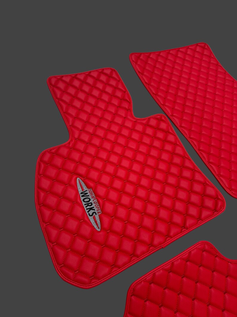 May include: Set of bright red car floor mats with a diamond-quilted pattern. The mats have a smooth, leather-like texture and feature the "John Cooper Works" logo in silver and white.