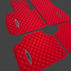 May include: Set of four bright red car floor mats with a diamond-quilted pattern. Each mat features the "John Cooper Works" logo in white, red, and gray. The mats are designed to fit a car's interior.