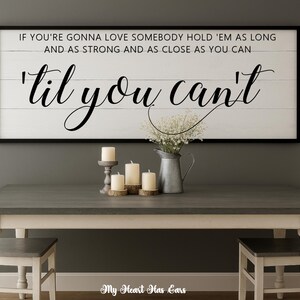May include: A framed wooden sign with the quote "IF YOU'RE GONNA LOVE SOMEBODY HOLD 'EM AS LONG AND AS STRONG AND AS CLOSE AS YOU CAN 'til you can't". The sign is above a gray table with candles and a vase.