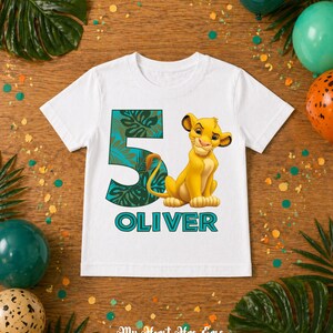 May include: White t-shirt featuring a cartoon lion cub and the number 5 in teal with a tropical leaf design. The name "OLIVER" is printed below the lion in teal. The shirt is decorated with balloons and confetti.