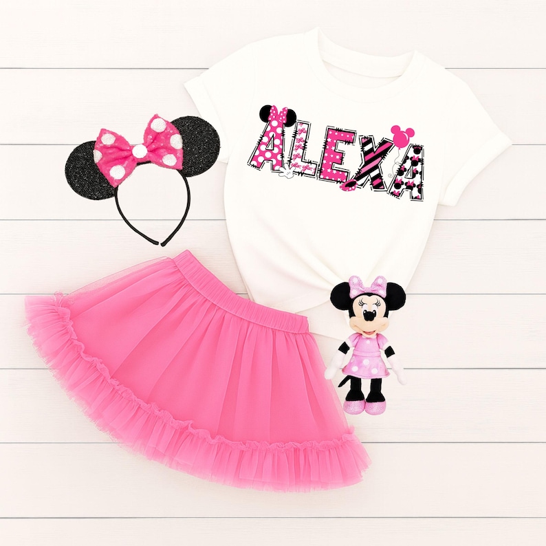 K&ouml;nnte beinhalten: Ein wei&szlig;es T-Shirt mit dem Namen "Alexa" in rosa und schwarz gemusterten Buchstaben, ein pinkfarbener T&uuml;llrock, ein Minnie Mouse-Haarreif und eine Minnie Mouse-Puppe. Das Outfit ist f&uuml;r ein Kind bestimmt.