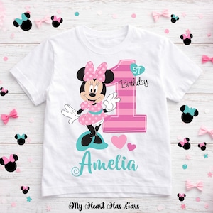 K&ouml;nnte beinhalten: Wei&szlig;es T-Shirt mit einem Minnie Mouse-Motiv in Rosa und Schwarz, mit der Zahl 1 und dem Text "1st Birthday". Der Name "Amelia" steht in t&uuml;rkisfarbener Schrift darunter. Dekorative Schleifen und Konfetti umgeben das Shirt.