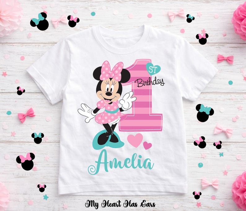 K&ouml;nnte beinhalten: Wei&szlig;es T-Shirt mit einem Minnie Mouse-Motiv in Rosa und Schwarz, mit der Zahl 1 und dem Text "1st Birthday". Der Name "Amelia" steht in t&uuml;rkisfarbener Schrift darunter. Dekorative Schleifen und Konfetti umgeben das Shirt.