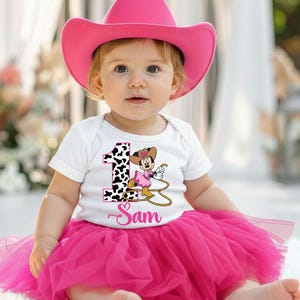 May include: A baby wearing a pink cowboy hat, white onesie with a cowgirl graphic, and a pink tutu skirt. The onesie has the number 1 with a cow print and the name "Sam" in cursive.
