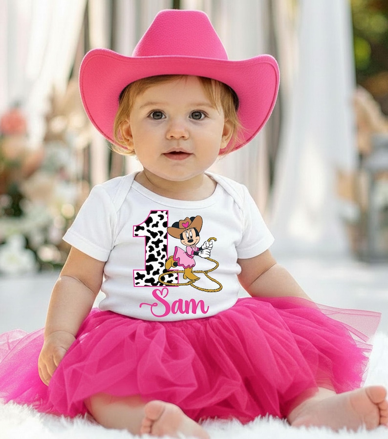 May include: A baby wearing a pink cowboy hat, white onesie with a cowgirl graphic, and a pink tutu skirt. The onesie has the number 1 with a cow print and the name "Sam" in cursive.