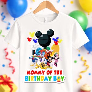 May include: A white short-sleeved t-shirt featuring a colorful graphic of various cartoon characters including Mickey Mouse, Minnie Mouse, Donald Duck, and Pluto. The text "MOMMY OF THE BIRTHDAY BOY" is printed in red, yellow, blue, and green letters.