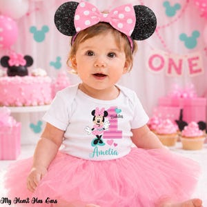 K&ouml;nnte beinhalten: Ein Baby-M&auml;dchen tr&auml;gt einen wei&szlig;en Body mit einem Minnie-Maus-Motiv und der Zahl "1" f&uuml;r ihren ersten Geburtstag. Sie tr&auml;gt einen rosa T&uuml;llrock und ein Minnie-Maus-Ohren-Stirnband mit einer rosa Schleife. Der Hintergrund umfasst rosa Dekorationen und einen Kuchen.