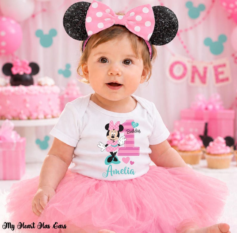 K&ouml;nnte beinhalten: Ein Baby-M&auml;dchen tr&auml;gt einen wei&szlig;en Body mit einem Minnie-Maus-Motiv und der Zahl "1" f&uuml;r ihren ersten Geburtstag. Sie tr&auml;gt einen rosa T&uuml;llrock und ein Minnie-Maus-Ohren-Stirnband mit einer rosa Schleife. Der Hintergrund umfasst rosa Dekorationen und einen Kuchen.