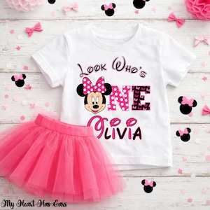 May include: A white t-shirt and pink tutu skirt set. The t-shirt features a Minnie Mouse graphic with the text "Look Who's ONE Olivia". The pink tutu is made of tulle. Decorative bows and confetti are scattered around the set.