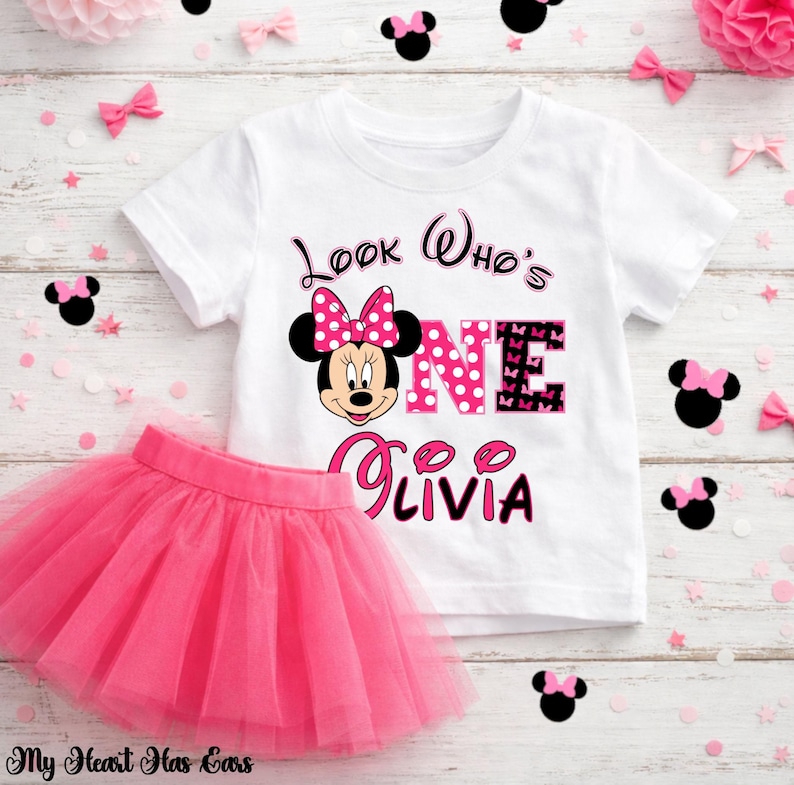 May include: A white t-shirt and pink tutu skirt set. The t-shirt features a Minnie Mouse graphic with the text "Look Who's ONE Olivia". The pink tutu is made of tulle. Decorative bows and confetti are scattered around the set.
