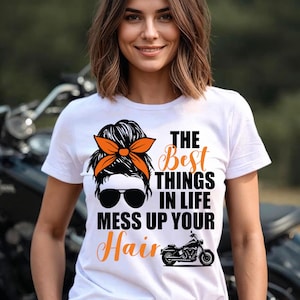 Puede incluir: Camiseta blanca con el texto "The Best Things in Life Mess Up Your Hair" en negro y naranja. El diseño incluye un gráfico de motocicleta y la cara de una mujer con gafas de sol y una diadema naranja.