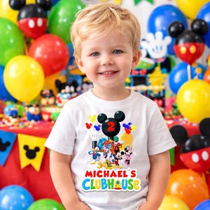 May include: A white short-sleeved t-shirt featuring a colorful graphic of cartoon characters including Mickey Mouse, Donald Duck, and Pluto. The design includes a large black Mickey Mouse ear icon with a red number 2 and the text Michael's Clubhouse.
