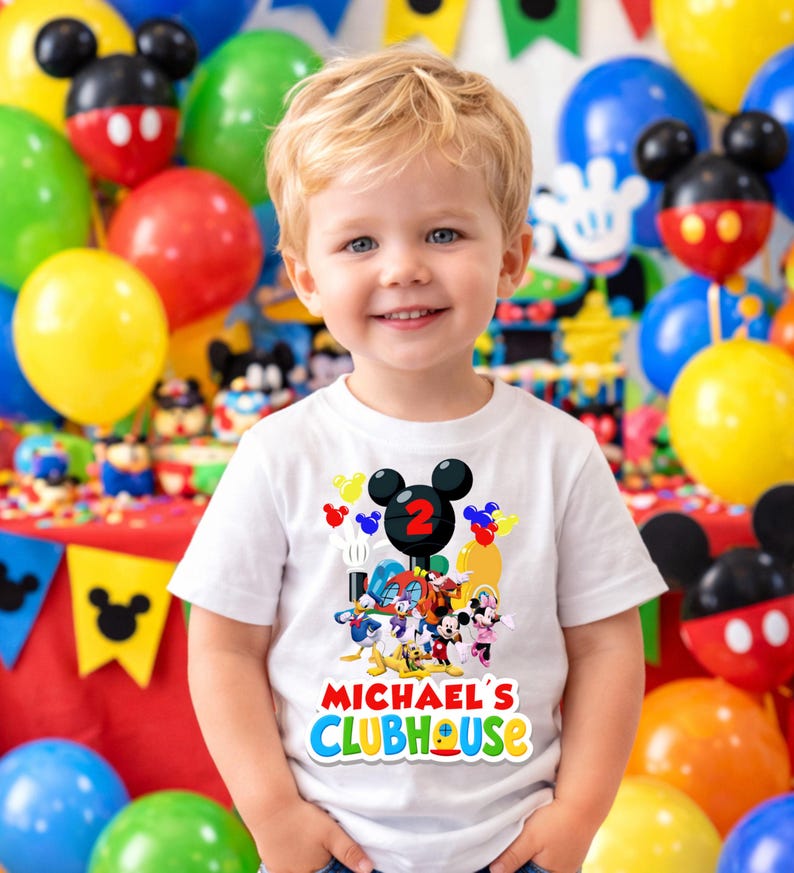 May include: A white short-sleeved t-shirt featuring a colorful graphic of cartoon characters including Mickey Mouse, Donald Duck, and Pluto. The design includes a large black Mickey Mouse ear icon with a red number 2 and the text Michael's Clubhouse.