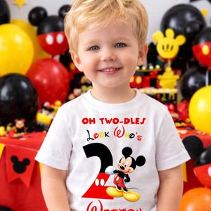 May include: A young child wearing a white t-shirt with a Mickey Mouse design. The shirt features the text "OH TWO-DLES Look Who's 2 Weston" in red and black. The background includes balloons and Mickey Mouse decorations.