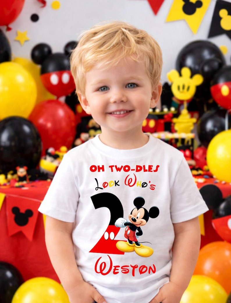 May include: A young child wearing a white t-shirt with a Mickey Mouse design. The shirt features the text "OH TWO-DLES Look Who's 2 Weston" in red and black. The background includes balloons and Mickey Mouse decorations.