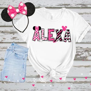 K&ouml;nnte beinhalten: Wei&szlig;es T-Shirt mit dem Namen "ALEXA" in rosa und schwarz gemusterten Buchstaben, mit Minnie-Mouse-Akzenten. Ein passendes rosa gepunktetes Schleifen-Stirnband und hellblaue Jeans sind ebenfalls im Bild.